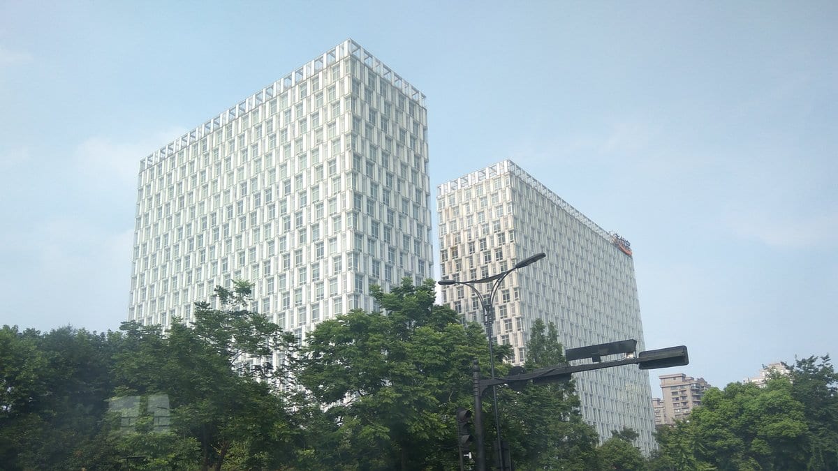 Two white office towers behind street trees, with a red Alipay sign near the roofline of the right tower.