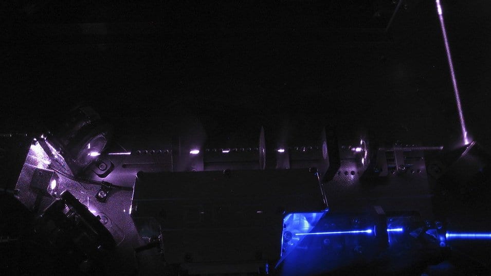 Violet and blue laser beams crossing mirror mounts and a frequency conversion cavity on a dark optical table.