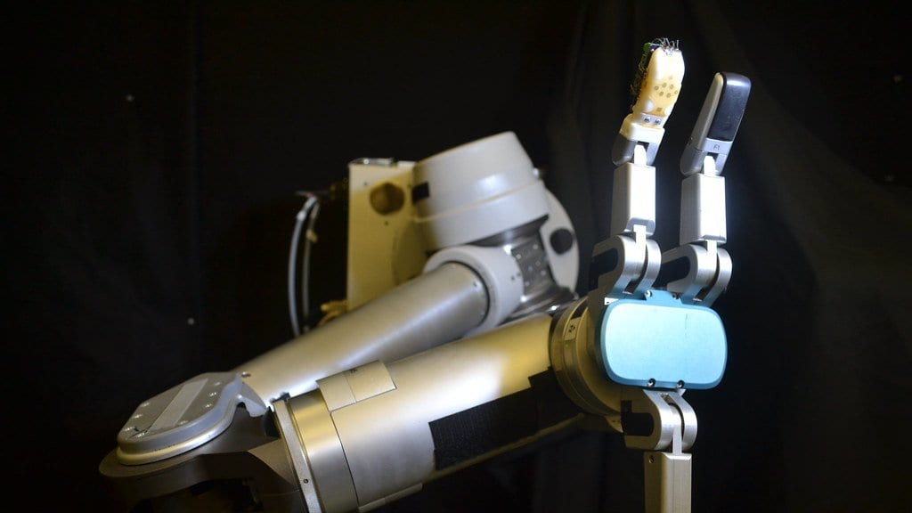 A metallic laboratory robot arm and gripper photographed against a black background, with a small pale sensing pad fitted over one fingertip.