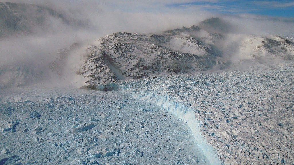 Aerial view of the calving front of Gyldenlove Glacier in southeast Greenland, a low wall of ice meeting a field of broken ice blocks, with cloud and dark mountains behind.