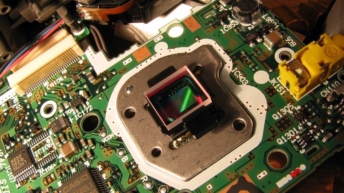 A camera's image sensor mounted at the center of a green circuit board inside an opened camera body, surrounded by ribbon cables and connectors.