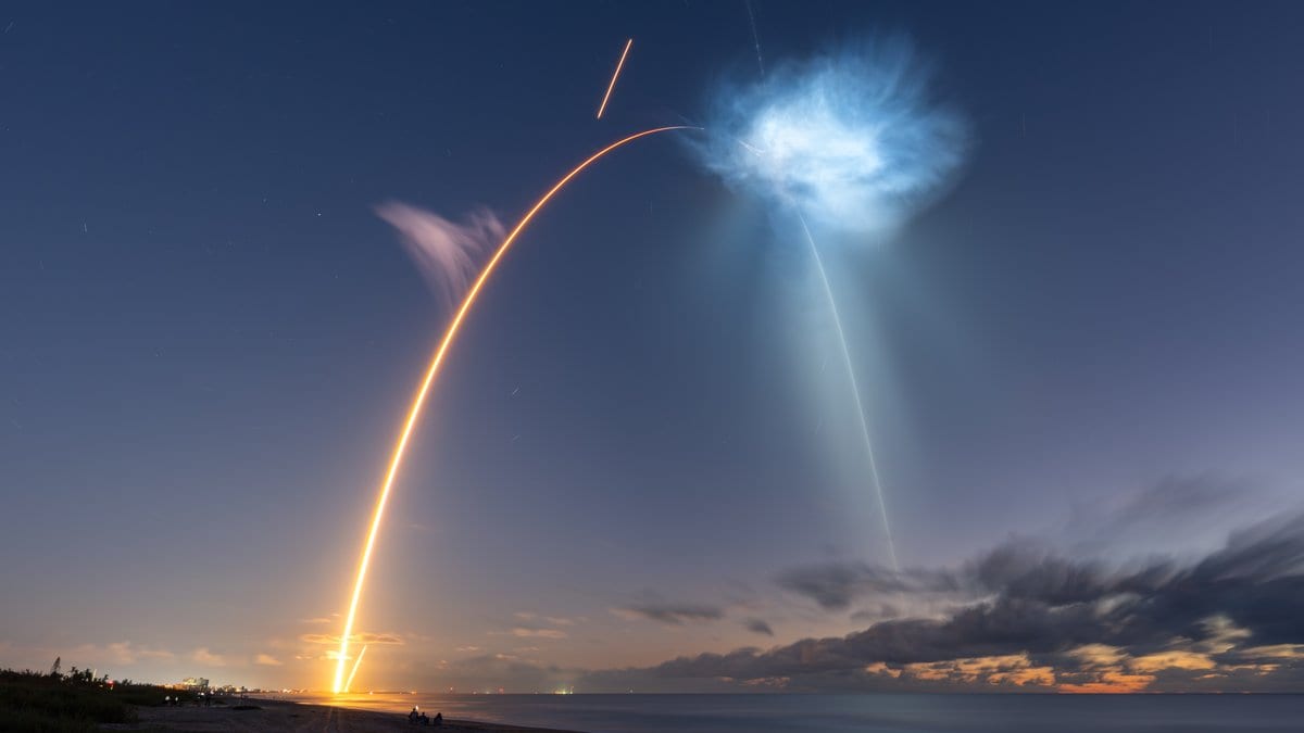 A long exposure shows the glowing orange arc of a Falcon 9 rocket rising from the Florida coast before sunrise, its upper-level exhaust plume lit pale blue high in the sky, with a few spectators on the dark beach below.