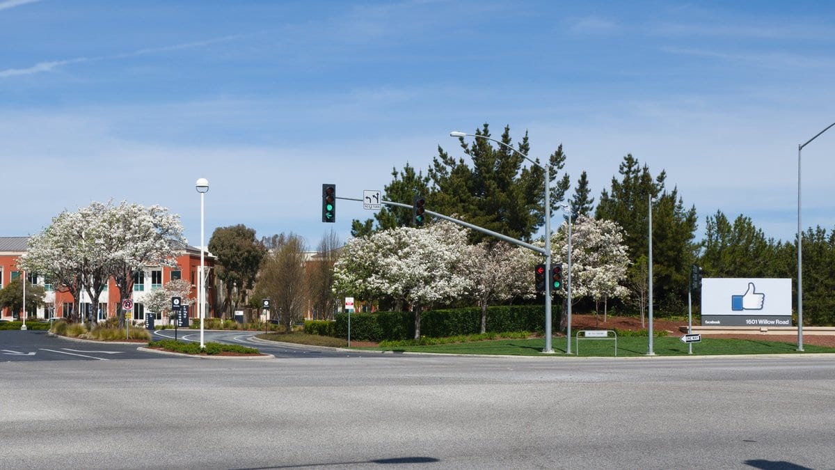 A roadside sign with a blue thumbs-up logo at the entrance to Meta's campus on Willow Road in Menlo Park, California, with office buildings and flowering trees behind it.