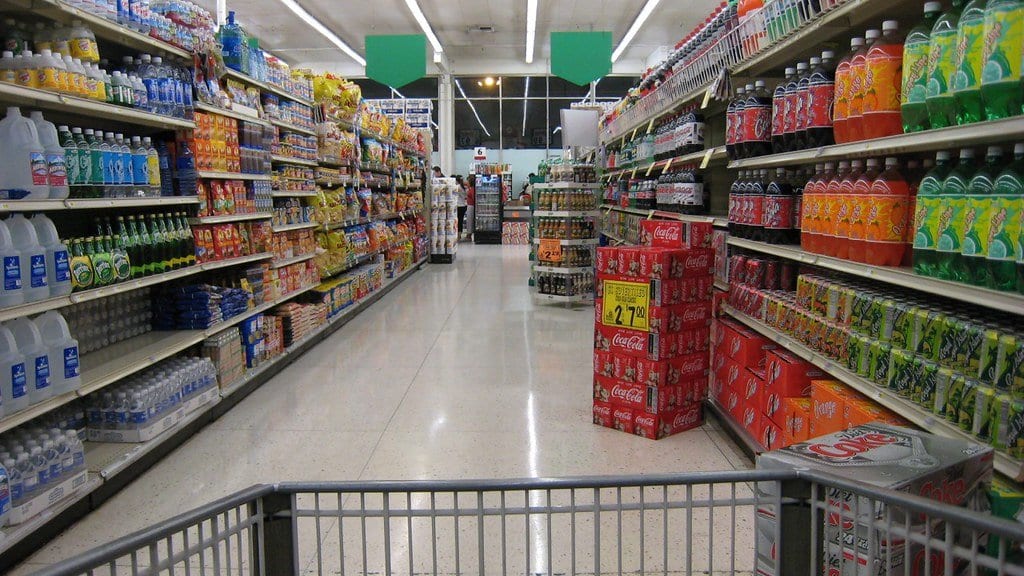 A supermarket aisle lined with bottled drinks, stacked soda cases and packaged snacks, seen from behind a shopping cart.