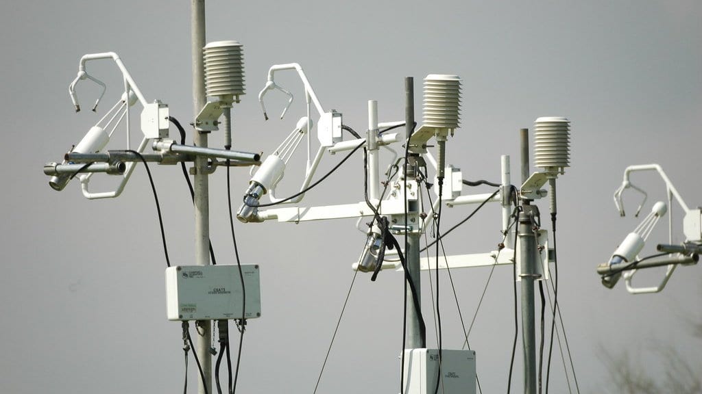Several metal masts carrying white sonic anemometers, radiation shields and instrument boxes stand against an overcast sky at a surface flux measurement site.