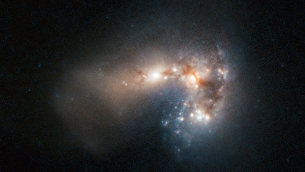 Hubble photograph of the compact starburst galaxy Haro 11: a bright, dust-laced core with clumps of blue-white young star clusters on one side, against a black sky.