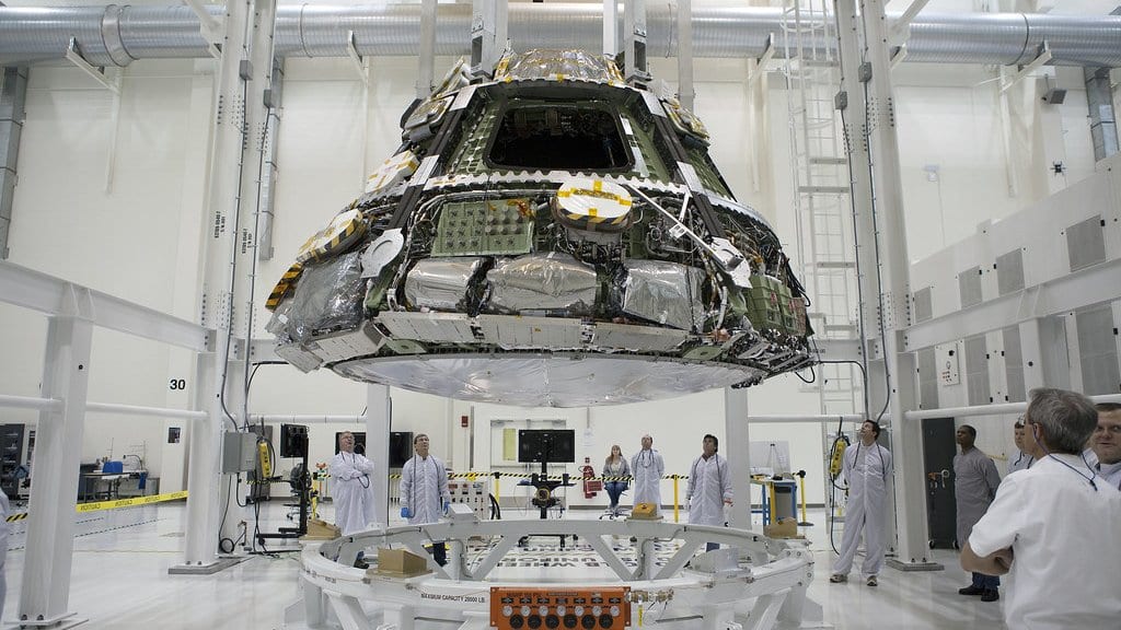 An Orion crew module hangs from overhead lifting gear above a white support stand while technicians in white coats watch from the floor of a high bay.