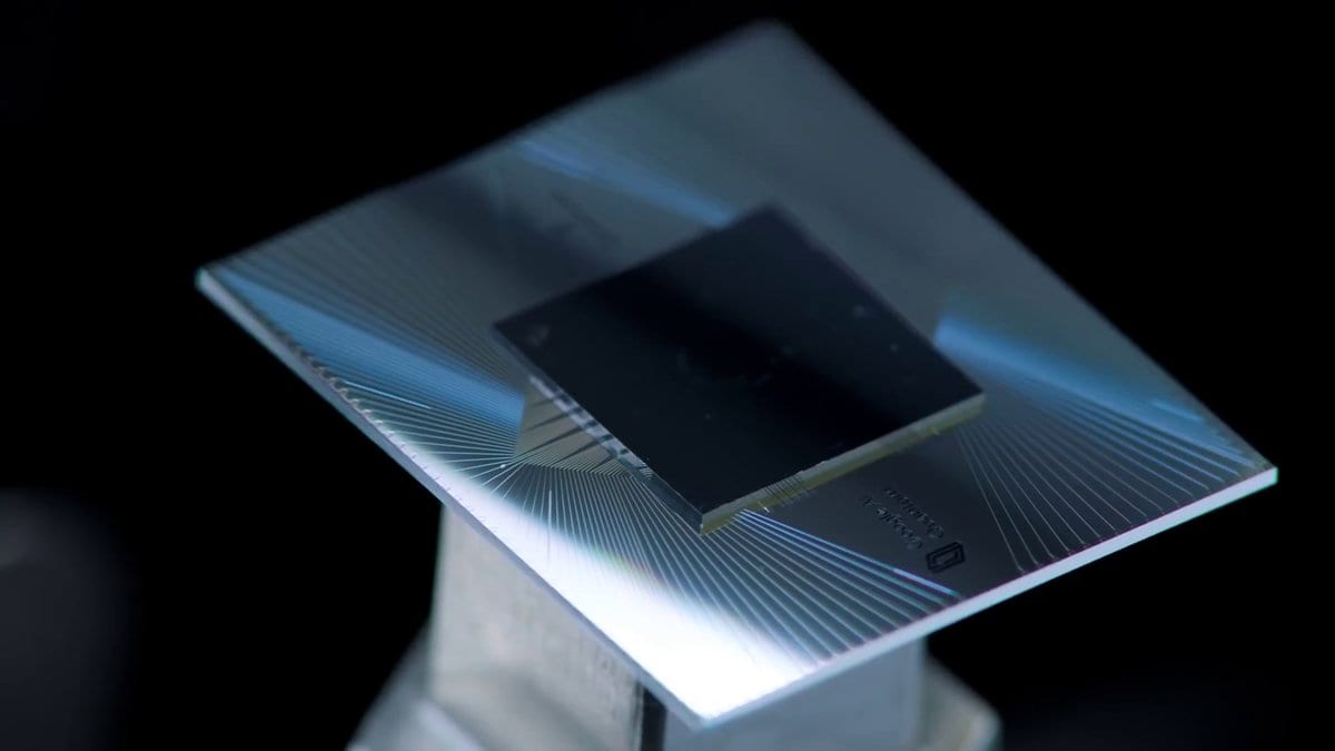 A dark square quantum computing chip mounted at the center of a square carrier, with fine metal traces fanning out from the chip toward the carrier's edges, against a black background.