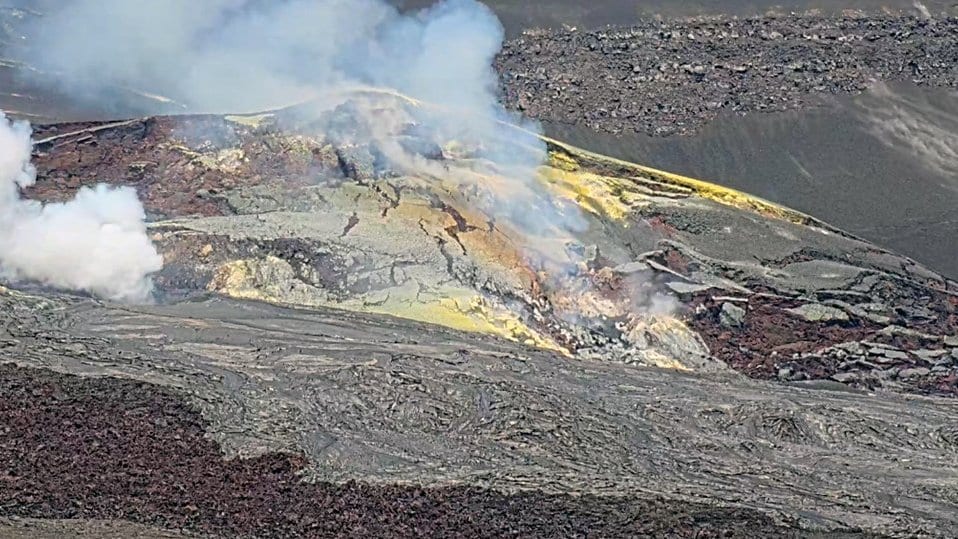 Steam rises from sulfur-stained ground on the dark lava floor of Halemaumau crater at the summit of Kilauea, with older gray lava and the crater walls behind.