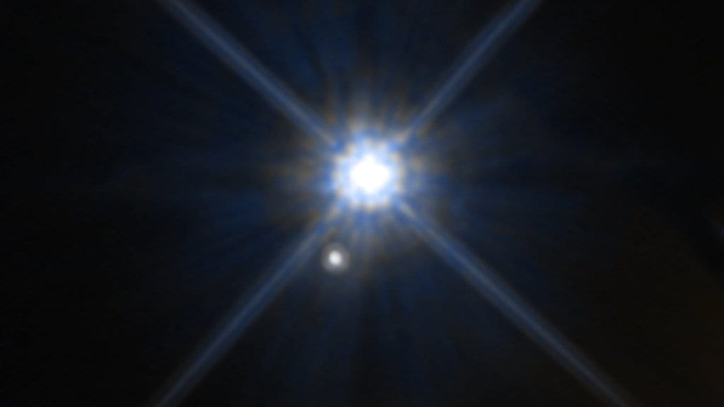 A Hubble Space Telescope image of the white dwarf Stein 2051 B as a bright white point with diffraction spikes, with a much fainter background star below it.