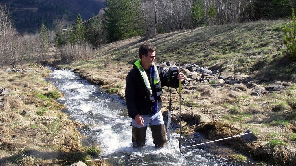 A hydrologist in waders stands in a shallow stream in a hilly valley, holding a flow meter on a wading rod, with a measuring tape stretched across the channel.