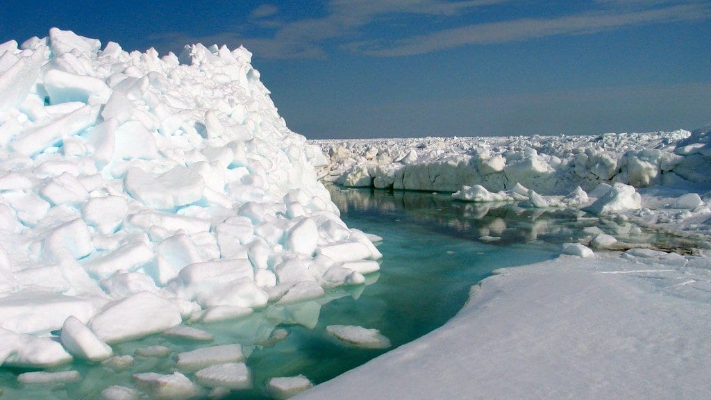 A ridge of broken sea ice blocks beside a narrow lane of open water in the Arctic pack ice, under a blue sky.