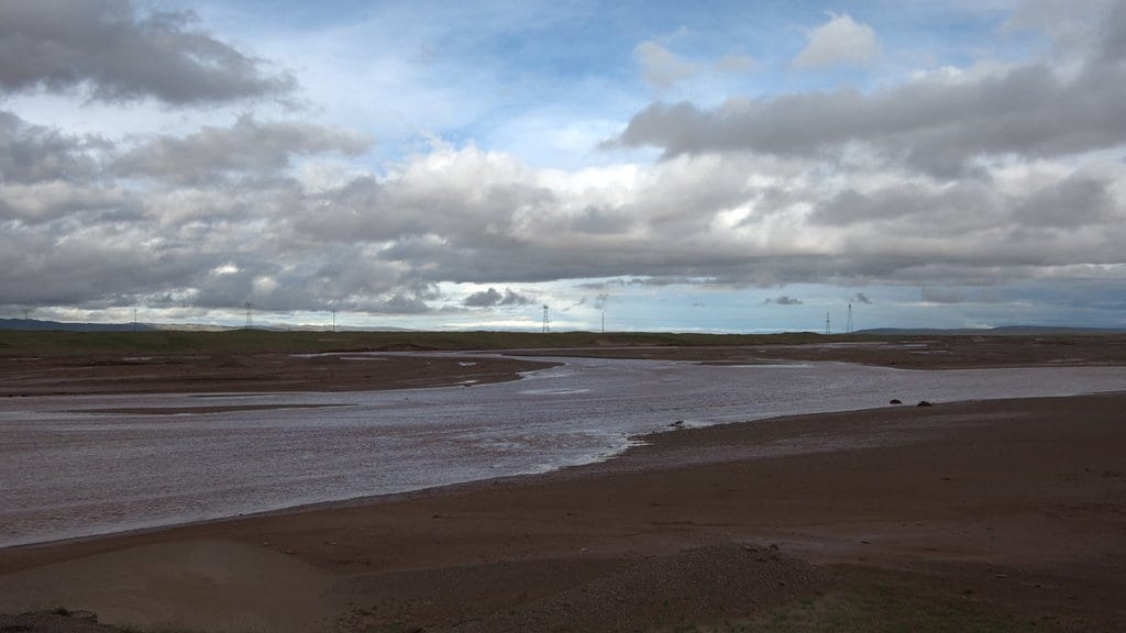 A braided river with pale sand and mud bars crossing flat plateau grassland under a cloudy sky.