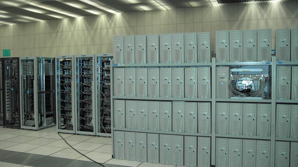 Rows of servers on open shelving and in cabinets in a large, brightly lit server hall.
