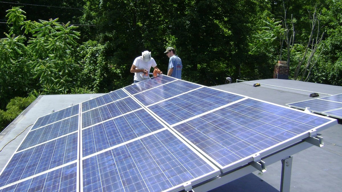 Two workers kneel beside a tilt-up photovoltaic array covering the flat roof of a house, with trees behind them.