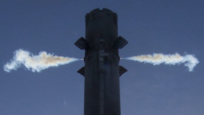 A rocket first stage against a deep blue sky, with two white jets of gas firing sideways from thrusters near the top of the booster.