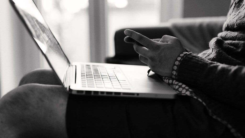 A person sits on a sofa with an open laptop resting on their lap, one hand reaching to the trackpad, in a black-and-white photograph of a sunlit room.