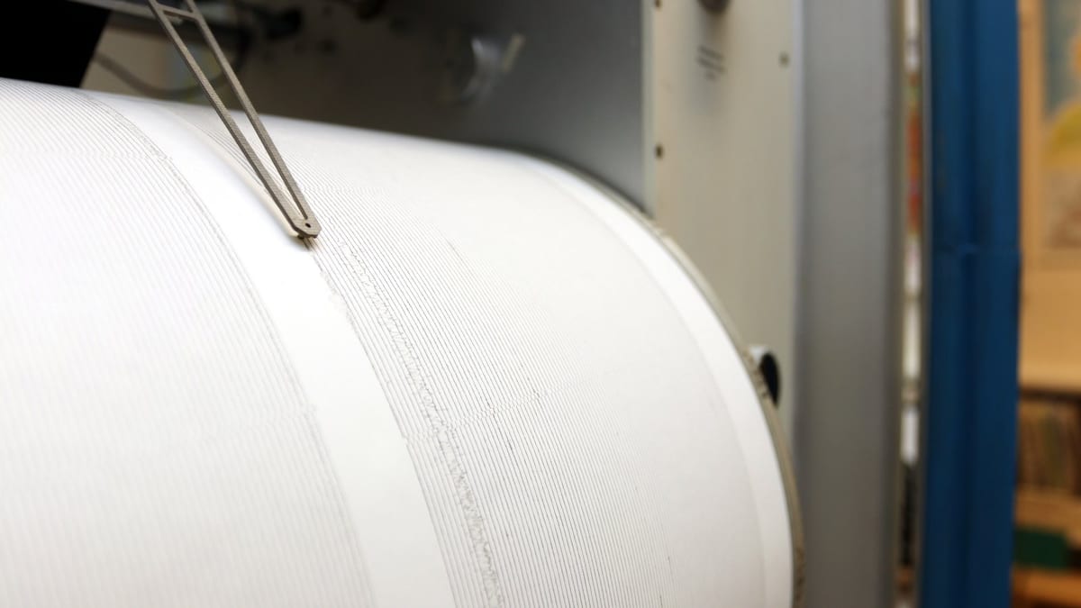Close view of a thin metal stylus resting on a white paper drum covered in fine horizontal trace lines, one band showing a short burst of wider wiggles