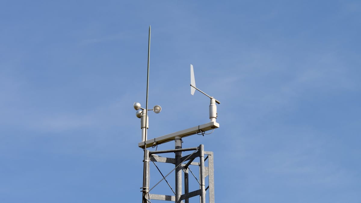 A galvanised lattice mast against a pale blue sky, topped by a cup anemometer, a wind vane on a short boom and a lightning rod