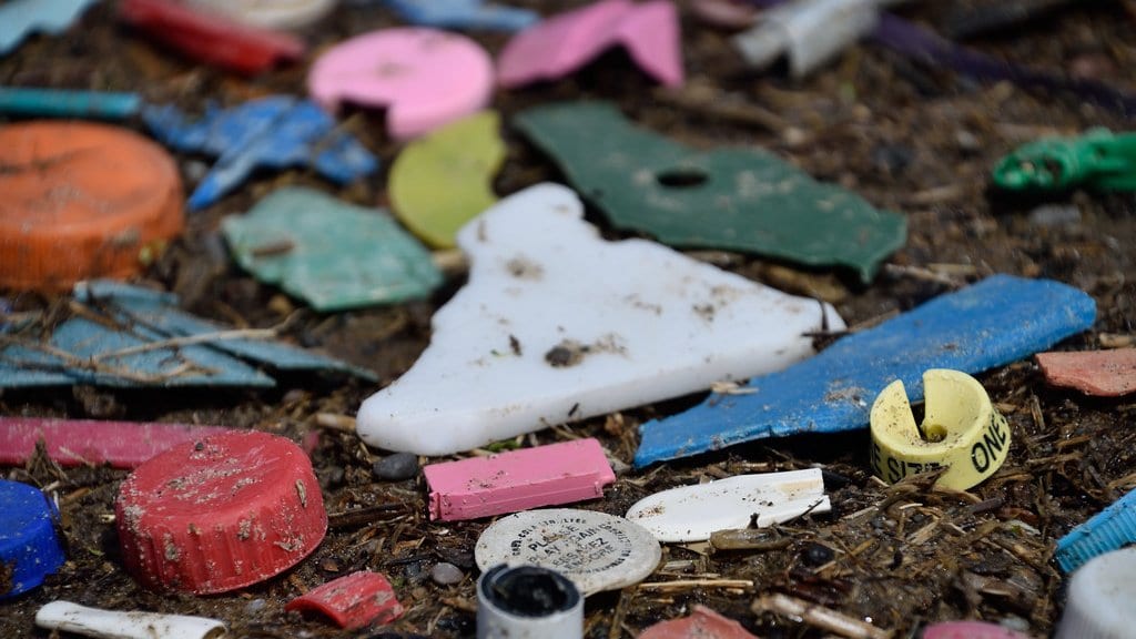Weathered fragments of colored plastic, bottle caps and broken pieces lying in sand and beach debris.