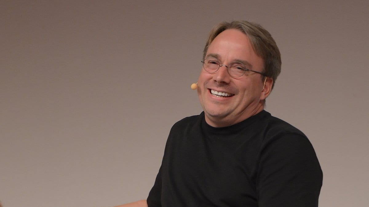 Linus Torvalds, in a black T-shirt and a headset microphone, smiles during a conference session.