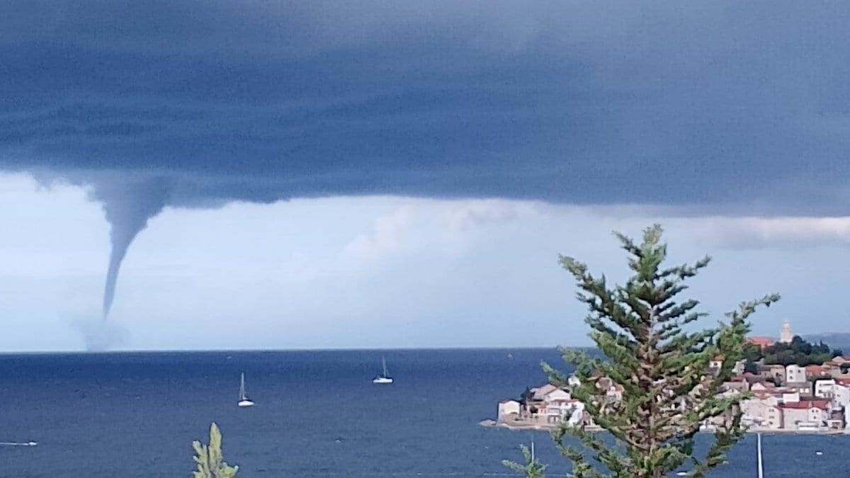 A narrow waterspout extends from the flat base of a dark storm cloud down to the sea, with sailboats and a coastal town to the right.