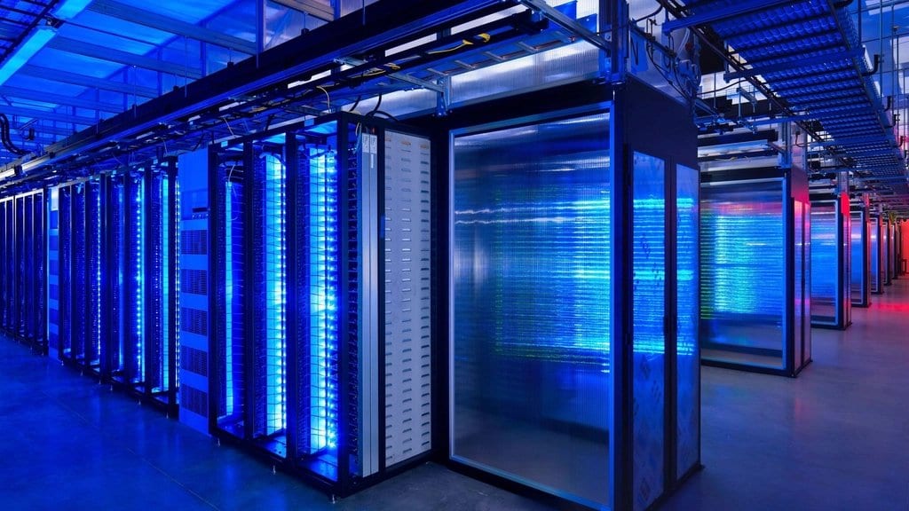 An aisle of server racks in a data center, lit blue by indicator lights.