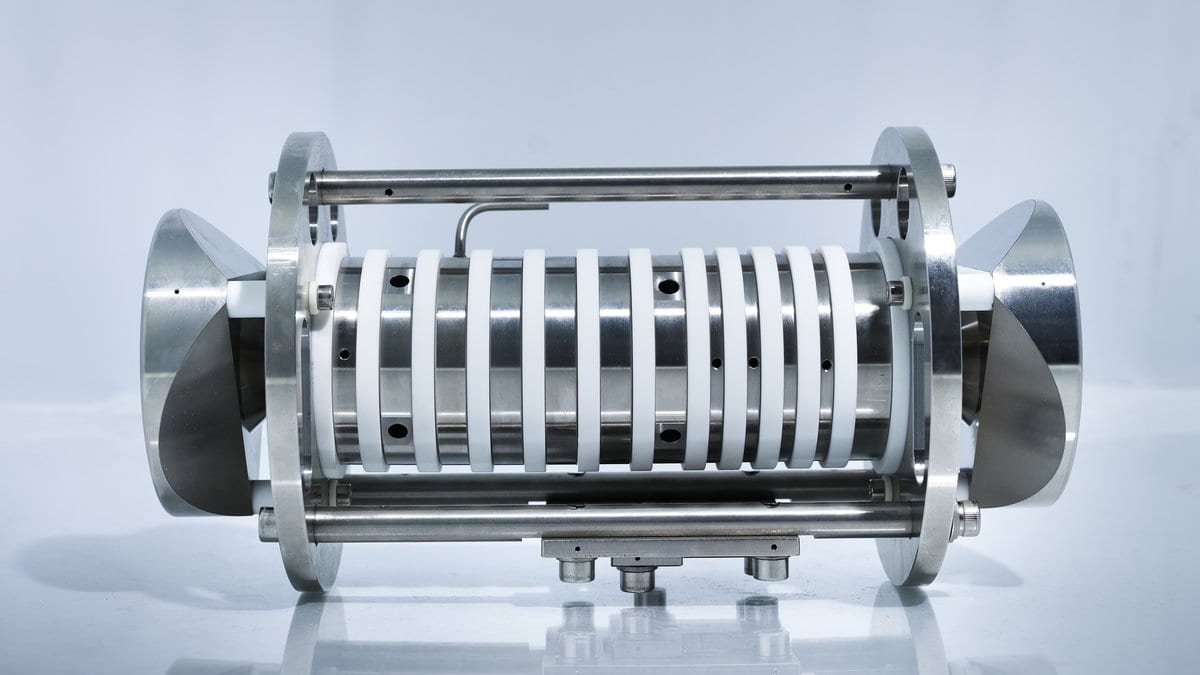 A cylindrical ion trap with stacked metal ring electrodes between two conical end caps, photographed against a pale background.