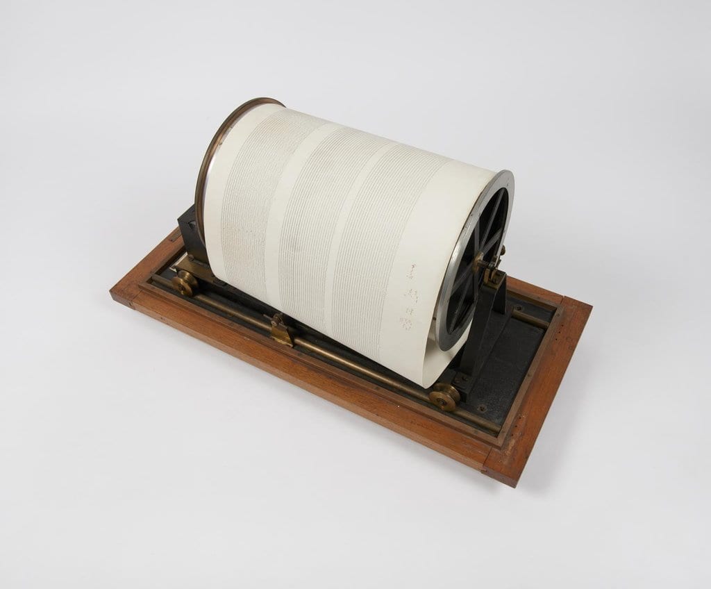 A cylindrical recording drum from an early seismograph, wrapped in paper marked with fine horizontal traces and mounted on a wooden base.