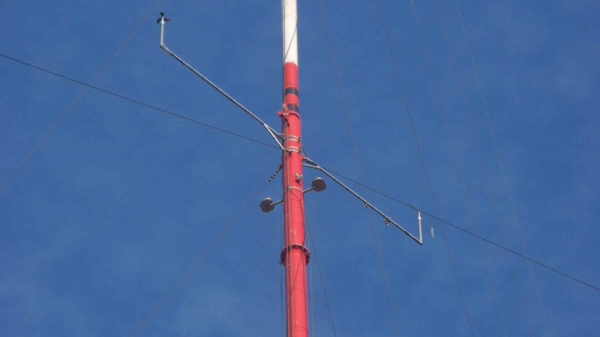 A red and white guyed mast against a deep blue sky, with booms carrying cup anemometers and a small wind vane