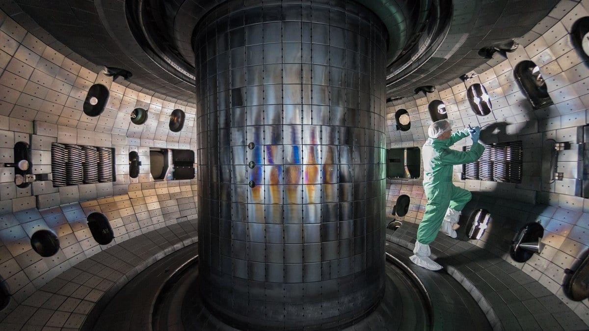 A technician in a protective suit stands inside the doughnut-shaped metal vacuum vessel of the DIII-D tokamak, beside its tiled central column.
