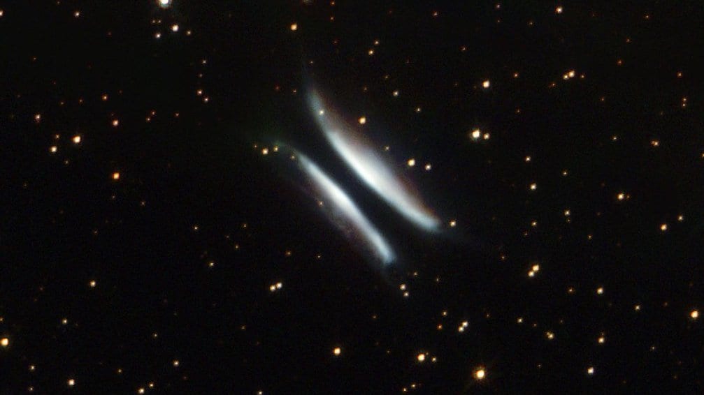 A narrow dark lane of dust runs between two curved, glowing lobes of scattered starlight, set against a dense field of stars.