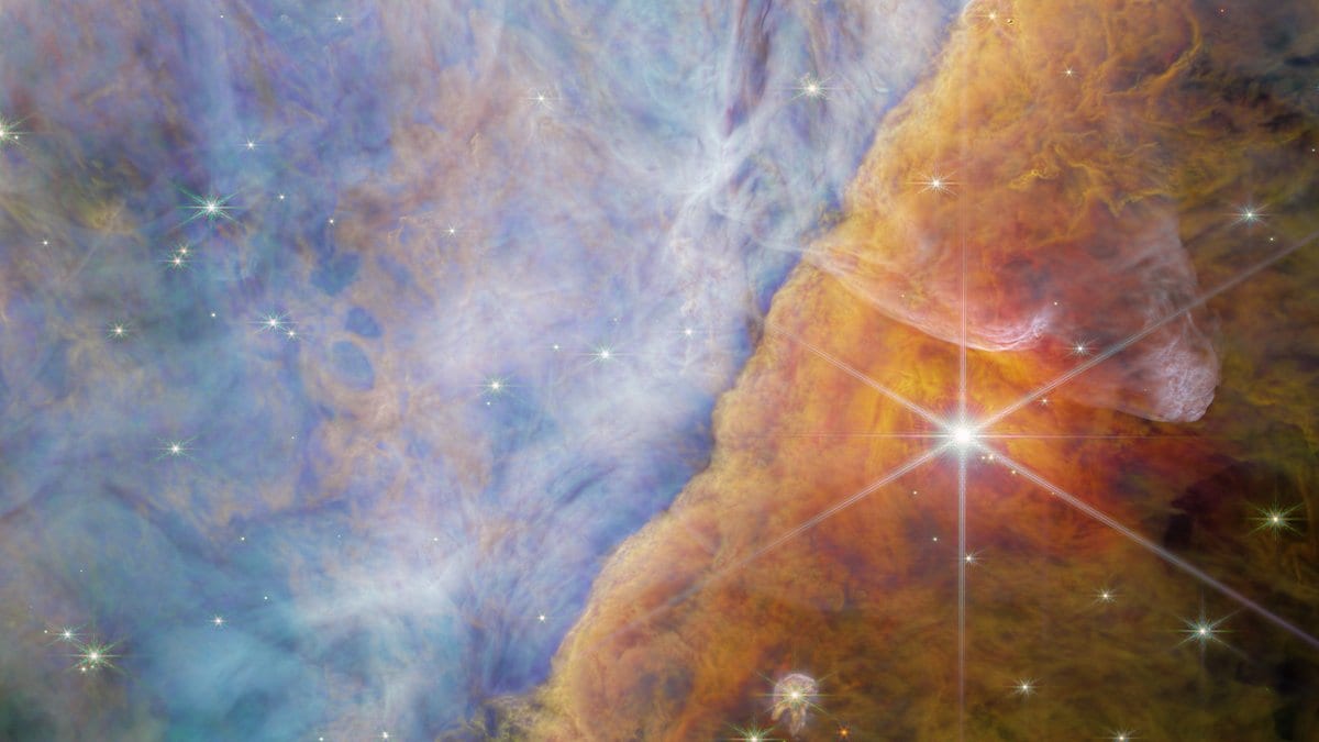 A near-infrared image of the Orion Bar, a glowing ridge of dense interstellar gas running diagonally across the frame, with wispy filaments along its irradiated edge and cooler blue-toned gas beyond it.