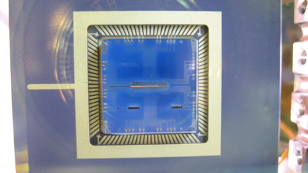 A square microfabricated ion trap chip with fine gold wire bonds, mounted in its package and photographed through the window of a vacuum chamber.
