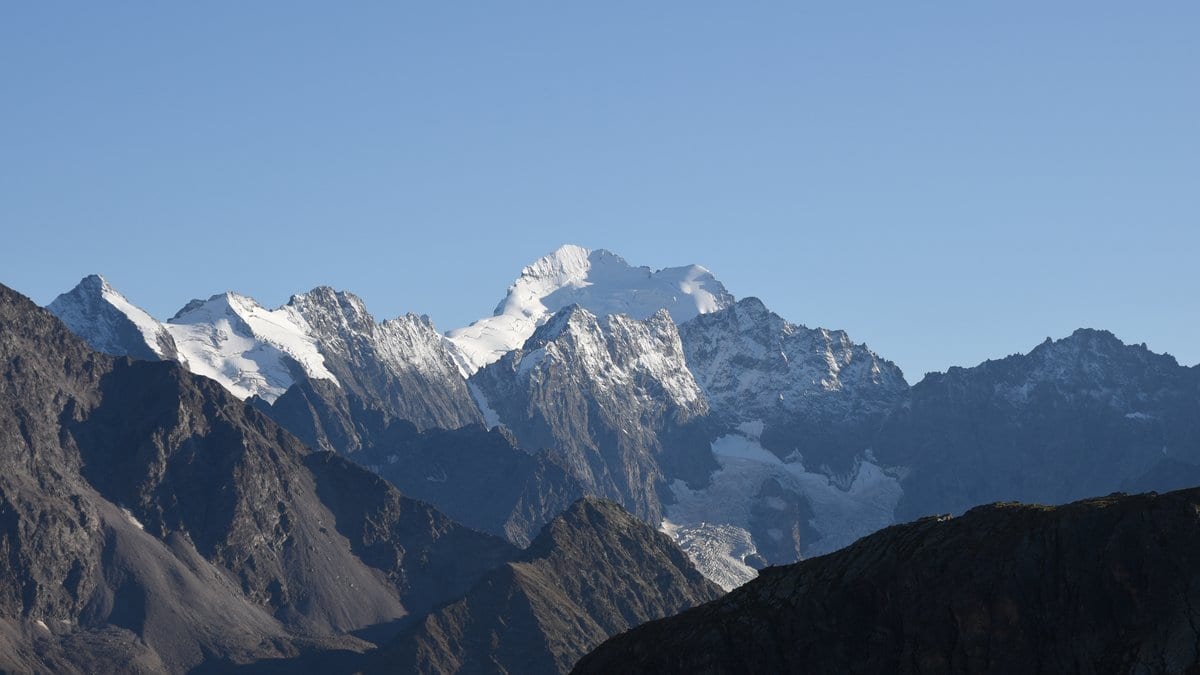 Snow-covered peaks of the Écrins massif in the French Alps under a clear sky, with dark rocky ridges in the foreground.