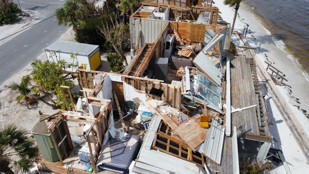 An aerial view of a beachfront house torn open to its framing, with lumber and sheet metal scattered across the sand beside the water.