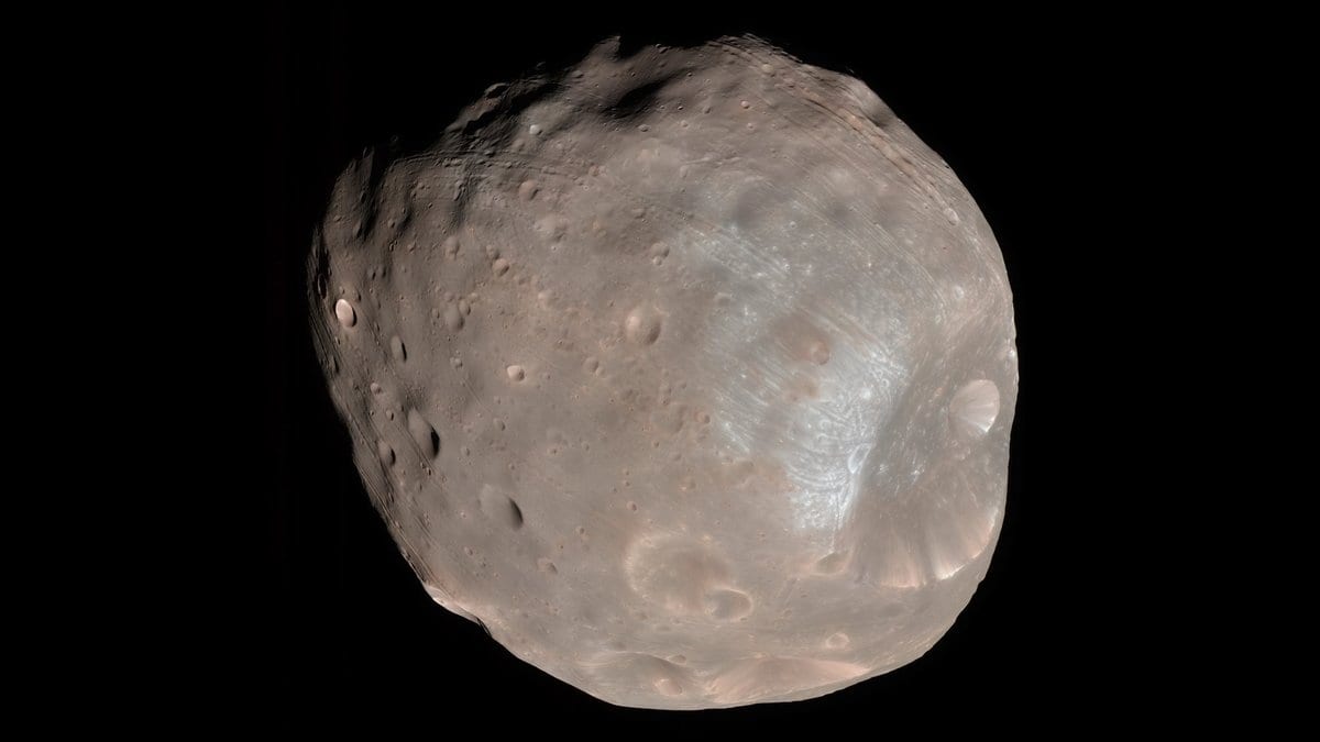 A color view of the same moon, its dusty surface pale pink and gray, with a bluish patch around the rim of its largest crater.