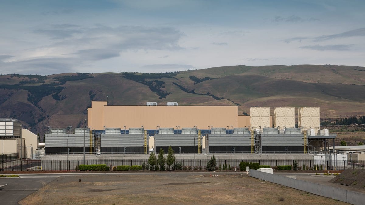 A long, low data center building with a row of cooling units along its side, set against dry hills under a cloudy sky.