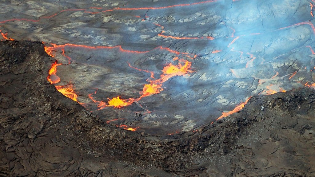 A crusted lava lake seen from above, bright orange cracks running across its gray surface, with a dark crater ledge in the foreground.