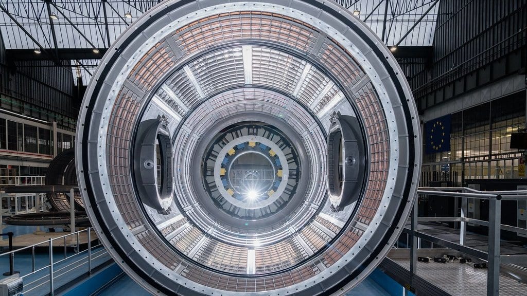 The open end of a cylindrical spacecraft module seen head-on inside a European integration hall, ribs and hatches receding toward a bright point at the far end.