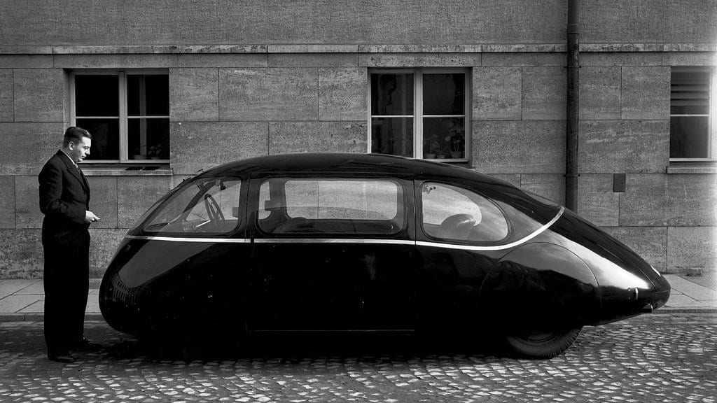 A black-and-white photograph of a teardrop-shaped experimental car parked beside a stone building, with a man in a suit standing at its nose.