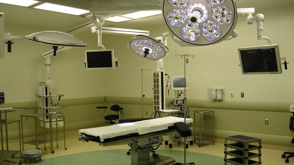 An empty operating room with a bare table at its center, two large multi-lamp surgical lights overhead, wall-mounted monitors and equipment booms around the walls.