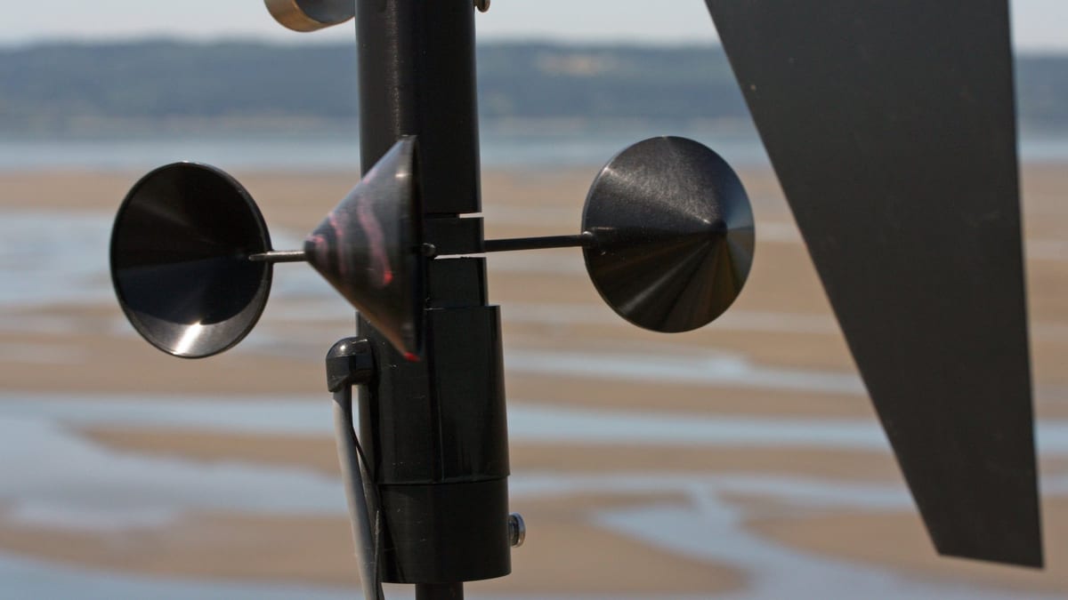 Close view of a black cup anemometer and the tail fin of a wind vane on a dark mast, pale tidal flats and a low shoreline out of focus behind