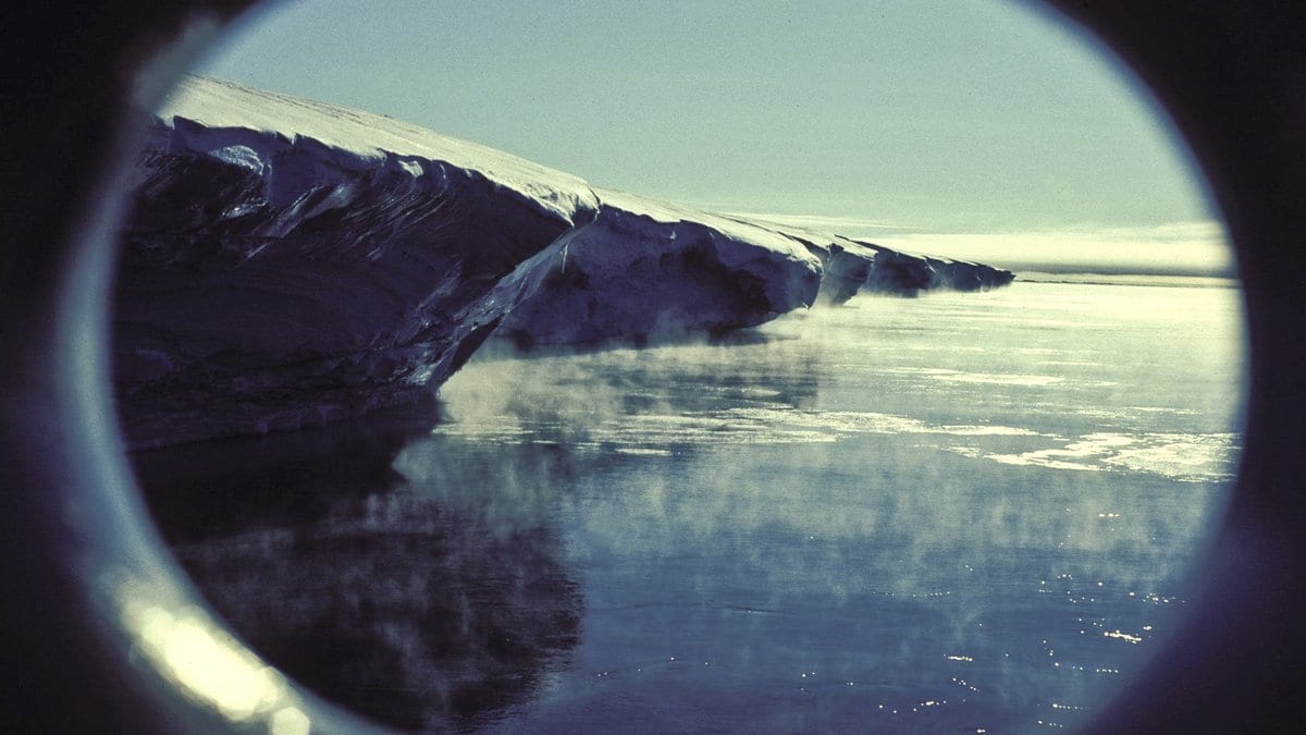 The floating cliff edge of an Antarctic ice shelf seen from a ship, with sea smoke rising off dark water at its foot.