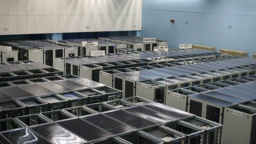 Rows of gray server cabinets fill a large computing room, with small overhead signs naming the services some of the racks carry.