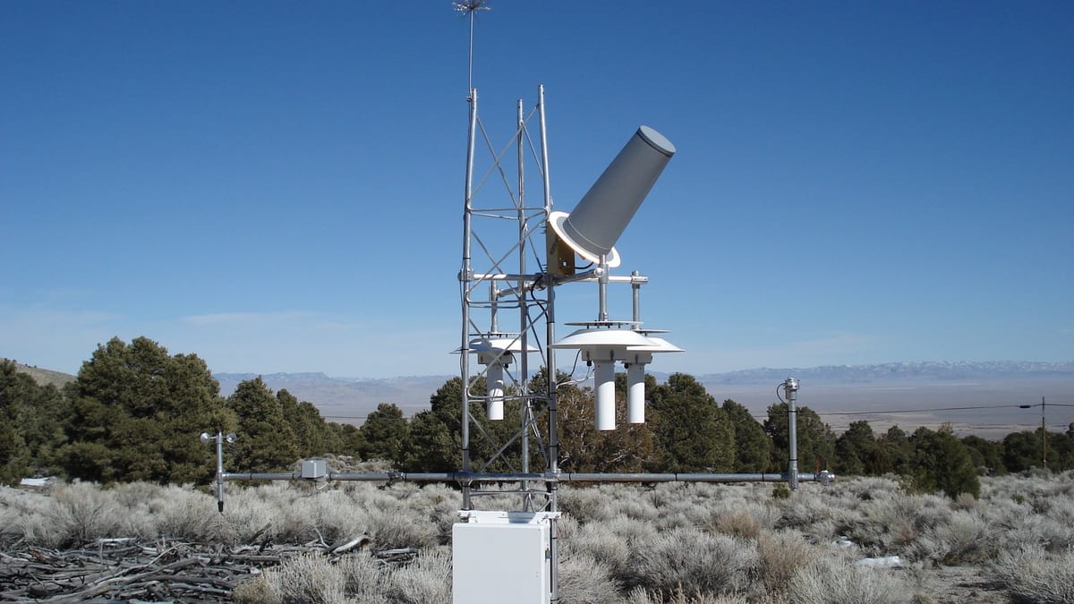 A lattice mast carrying shielded temperature sensors, a tilted radiation sensor and a wind gauge on a boom, standing in sagebrush with juniper and distant mountains under a deep blue sky