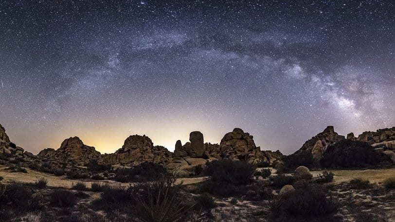 A wide arc of stars spans a dark sky above a rocky desert floor scattered with boulders and low brush.