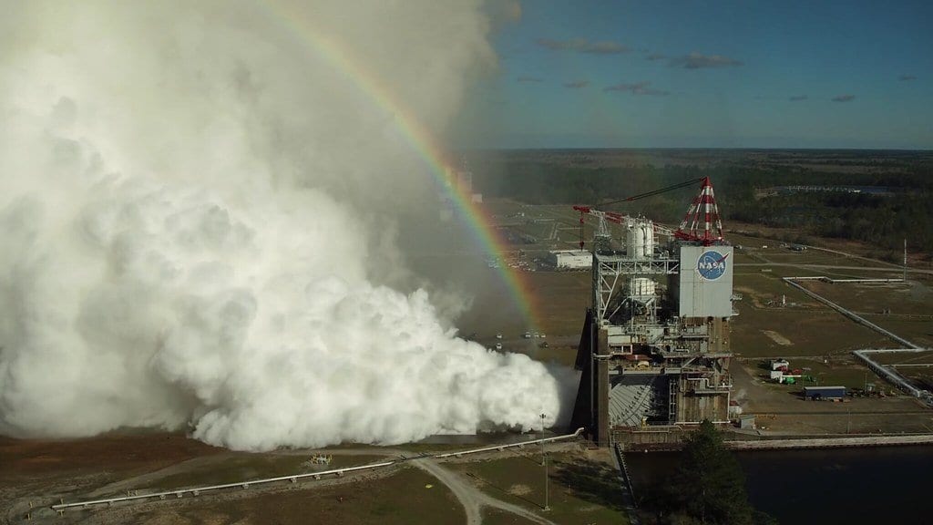 A rocket engine test stand releasing a huge white steam cloud across the ground, with a rainbow arcing above it under a blue sky.