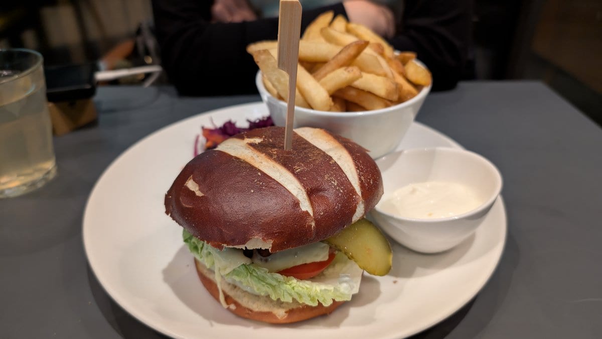 A plant-based burger in a pretzel bun on a white plate, with a bowl of fries and a pot of dip behind it.