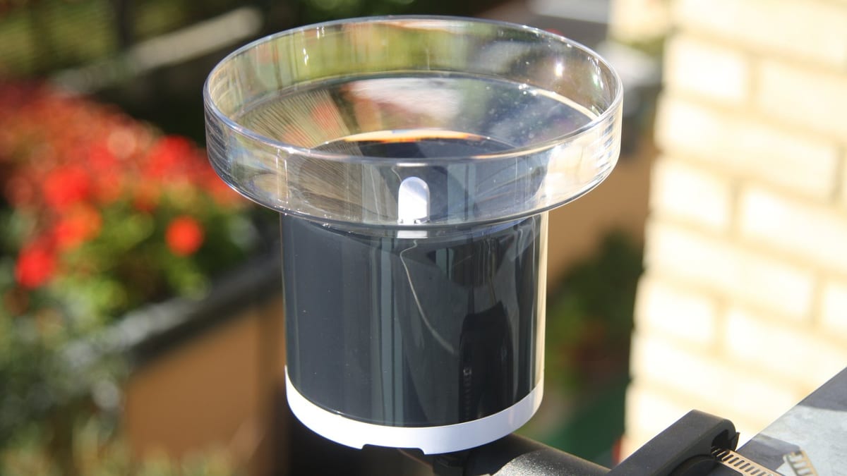 A clear plastic rain gauge funnel on a dark cylinder clamped to a metal rail, red flowers and a pale brick wall out of focus behind